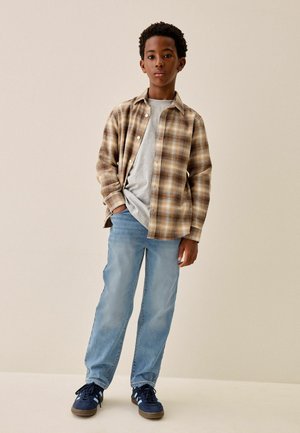 Dreng stående med den ene hånd i lommen iført en brun ternet skjorte over en grå t-shirt, lyseblå jeans og marineblå sneakers.