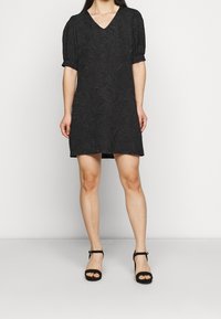 Mustan lyhyen hihansuiden mekon, jossa on V-muotoinen kaula-aukko ja teksturoitu lehtikuvio, rento istuvuus ja hieman levenevä siluetti.