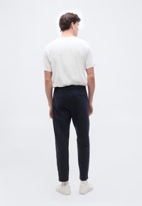 Άνδρας που στέκεται πλάτη, φοράει λευκό μπλουζάκι, μαύρο παντελόνι, λευκές κάλτσες και λευκά αθλητικά παπούτσια σε απλό λευκό φόντο.