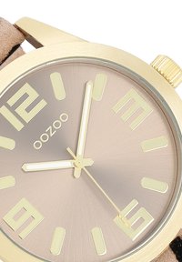 Gold-tone watch with a round face, beige dial, large hour markers, and three hands. Features a textured beige strap and brand logo.