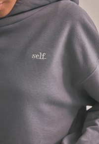 Szara bluza z kapturem wykonana z miękkiego materiału, z białym haftem na przodzie przedstawiającym słowo "self." o luźnym kroju i gładkiej teksturze.