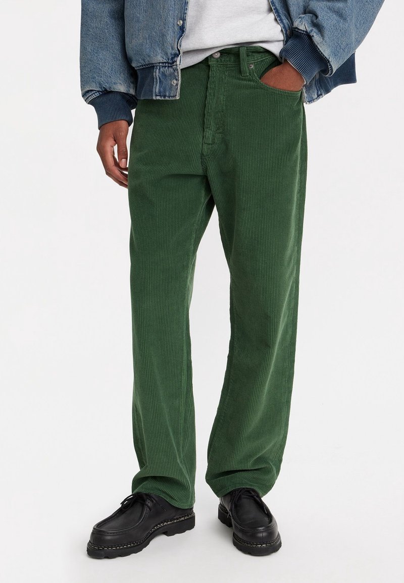 Personne portant un pantalon en velours côtelé vert avec une main dans la poche, des chaussures noires, une chemise blanche et une veste en jean bleue sur fond blanc.