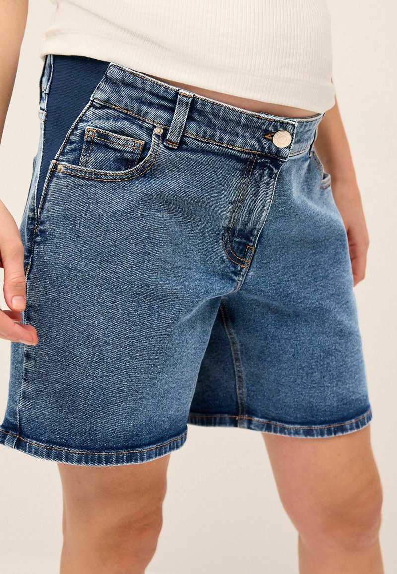 Short en denim bleu mi-long avec des panneaux latéraux élastiques porté par une personne vêtue d'un haut blanc sur un fond uni.