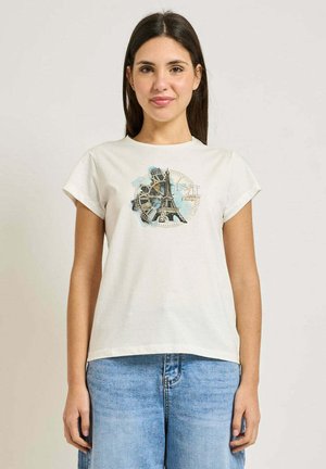 Giovane donna che indossa una maglietta bianca con disegno della Torre Eiffel e un orologio, abbinata a jeans azzurri, in piedi davanti a uno sfondo semplice.
