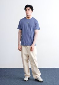 Nike blå bomull T-skjorte med grønn logo, kombinert med beige løse bukser og grå sneakers. Modellen står mot en enkel bakgrunn.