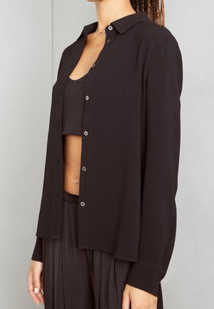 Schwarzes, geripptes Langarmhemd mit Kragen, Frontknöpfen und lässiger Passform. Getragen über einem passenden schwarzen Cropped Top und einer Hose.