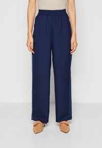 Pantalons larges navy avec une taille élastique, présentant un tissu lisse et des accents en satin sur les côtés. Assortis à des ballerines beiges à bout pointu.