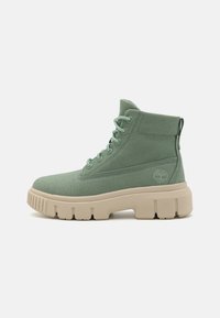 Stivaletti alla caviglia in tela verde con suola beige spessa, caratterizzati da un design con lacci, un colletto imbottito e dettagli con logo sul lato.