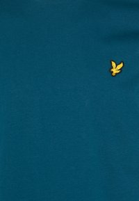 Camiseta de algodón en color verde azulado con cuello redondo, que presenta un pequeño logotipo de un pájaro amarillo bordado en el lado izquierdo del pecho. Textura suave, mangas cortas.