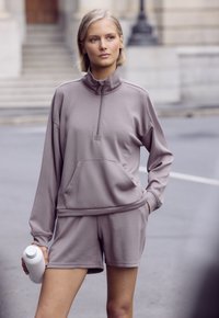 Gaiši purpura sporta komplekts, kurā ietilpst ar rāvējslēdzēju, augstu apkakli un garām piedurknēm topi ar priekšējo kabatu un atbilstošas šortus, izgatavots no gludas auduma.