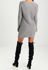 Kvinna sedd bakifrån i en grå stickad klänning och svarta ankelhöga stövlar i mocka, stående på ett enkelt vitt golv.