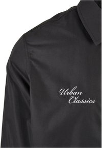 Camisa negra con el logotipo bordado "Urban Classics". Presenta un collar clásico y una textura suave. Diseño simple y moderno sin patrones.