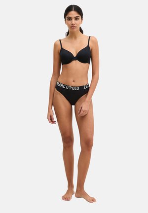 Kobieta stoi boso, ubrana w czarny biustonosz i czarne majtki z logo na pasku, na tle jasnego, jednolitego tła.