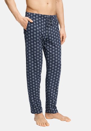 Uomo che indossa pantaloni del pigiama blu navy con motivi geometrici bianchi, in piedi a piedi nudi con una mano in tasca su sfondo uniforme.