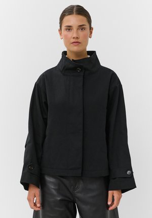 Femme portant une veste noire à col montant avec des poignets boutonnés et un pantalon en cuir noir, debout devant un fond blanc uni.
