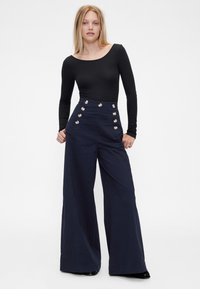 Marineblå bukser med høyt liv og vide ben, med sølvknapper foran, laget av et glatt stoff. Kombinert med en svart langermet topp.