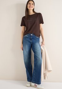 Femme souriante, portant un t-shirt marron foncé avec un texte, un jean large bleu, des baskets blanches, tenant une veste beige clair sur un fond uni.