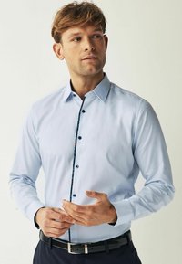 Helehelesin sinine särk, mis on valmistatud tekstureeritud kangast, omab klassikalist kraed ja kontrastset mustrit nööpide plaanis.