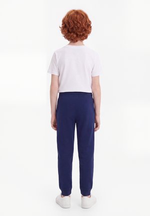 Enfant aux cheveux roux bouclés, portant un t-shirt blanc, un pantalon bleu marine et des chaussures blanches, debout de dos sur un fond blanc uni.