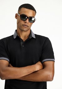 Schwarzes Poloshirt mit grauen Akzenten am Kragen und an den Ärmeln, das einen gepunkteten Innenspiegel hat. Das Model trägt dunkle Sonnenbrillen.