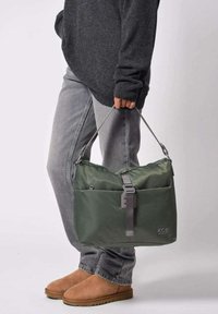 Bolso de hombro de nylon verde con correa ajustable, diseño elegante, amplio compartimento principal, acentos grises, logotipo en el frente, llevado por una persona.