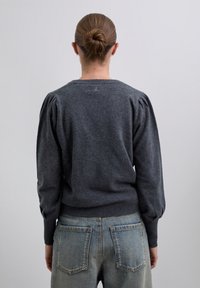 Femme avec un chignon portant un pull gris foncé à manches longues bouffantes et un jean taille haute bleu clair, se tenant devant un fond uni.