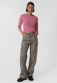Top corto de manga larga rosa combinado con pantalones de pierna ancha beiges con un estampado de leopardo negro. Modelo de pie con las manos en los bolsillos. Calzado negro.