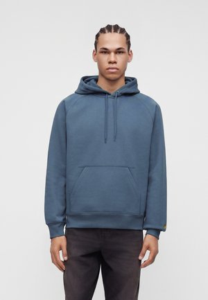 Jeune homme aux cheveux tressés portant un sweat à capuche bleu et un pantalon noir, debout devant un fond clair uni.