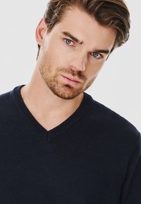 Granatowy sweter z dekoltem w kształcie litery V, wykonany z miękkiej tkaniny, z teksturowanym splotem i subtelnym detalem szwu przy dekolcie, noszony przez modela.