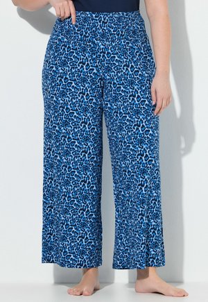 Personne portant un pantalon bleu imprimé léopard ample, longueur cheville avec une taille élastique, debout pieds nus sur fond blanc.