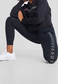 Persoon knielend in een zwart Calvin Klein Performance sweatshirt en leggings met witte logo, en witte sportschoenen op een grijze vloer.