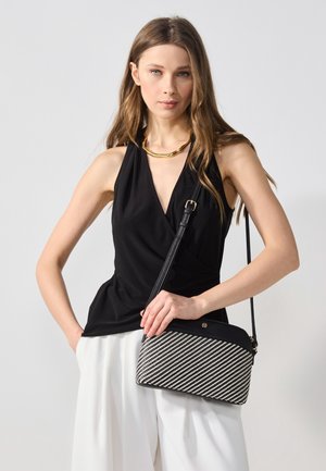 Kobieta ubrana w czarny bezrękawnik, białe spodnie, złoty naszyjnik, trzymająca czarno-białą torebkę na ramię na jednolitym tle.