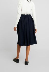 Marineblå midi-nederdel med en samlet talje, lavet af blødt stof. Parret med en hvid striktrøje og sorte slip-on sko.