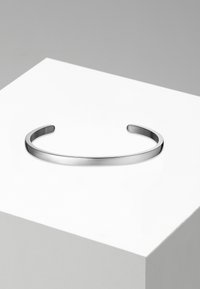 Bracelet en argent avec une surface lisse et polie, présentant une forme minimaliste et des extrémités ouvertes, exposé sur un piédestal blanc.