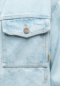 Φωτεινό μπλε τζιν μπουφάν με τσέπη που κλείνει με καπάκι και στρογγυλό μεταλλικό κουμπί με χαραγμένο το λογότυπο Timberland.