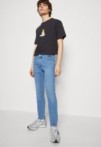Camiseta negra con estampado gráfico, jeans azul claro y zapatillas grises. El modelo posa frente a un fondo neutro, mostrando un atuendo casual.