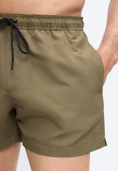 Άνδρας που φοράει χακί πράσινα μαγιό με μαύρο κορδόνι, το δεξί του χέρι στην τσέπη, δείχνοντας το κάτω μέρος του κορμού και τα πάνω μέρη των ποδιών του πάνω σε λευκό φόντο.