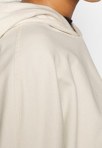 Sudadera beige con capucha, con una textura suave, un ajuste ligeramente holgado y detalles visibles de costuras a lo largo de las mangas y el cuerpo.