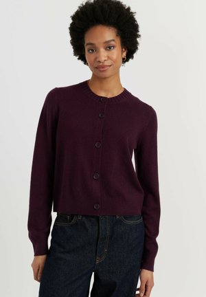 Femme aux cheveux bouclés portant un cardigan boutonné violet foncé et un jean bleu foncé, debout devant un fond uni.