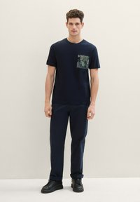 Camiseta de manga corta azul marino con tejido texturizado y bolsillo con un patrón verde, combinada con pantalones oscuros y zapatillas negras. Diseño simple y casual.