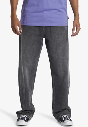 Baggy dark gray denim jeans with a faded texture and purple logo detail on the left front pocket. Worn with black and white sneakers.