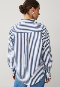 Kvinde med hår i en knold iført en blå og hvid vertikalt stribet skjorte med knapper og blå jeans, vendende væk mod en ensfarvet baggrund.
