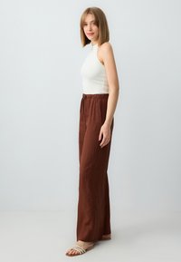Pantaloni ampi in lino marrone con vita regolabile, abbinati a un top senza maniche bianco a costine e sandali flat con cinturini.