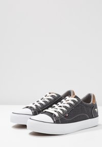 Černé plátěné tenisky s bílou gumovou podrážkou, kulatou špičkou a béžovými akcenty. Obsahují bílé tkaničky a švy, texturovanou látku a detail loga.