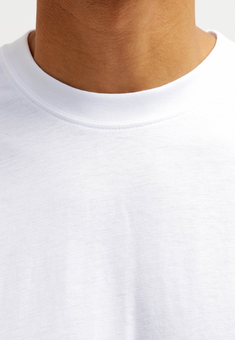Primo piano del collo e della parte superiore del petto di una persona che indossa una t-shirt bianca a girocollo semplice su uno sfondo bianco.
