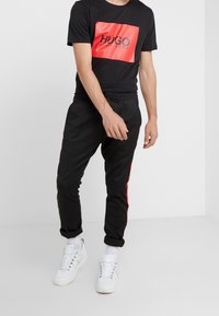 Camiseta negra con un logotipo rectangular rojo y texto negro; pantalones negros con franjas laterales rojas; zapatillas blancas; corte relajado.