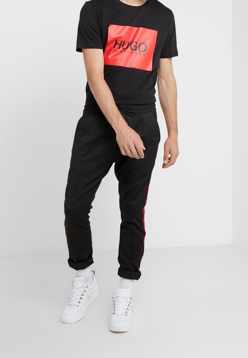 Camiseta negra con un logotipo rectangular rojo y texto negro; pantalones negros con franjas laterales rojas; zapatillas blancas; corte relajado.