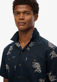 Camisa de manga corta en color marino con bordado de dragón plateado, dos bolsillos delanteros y textura similar a la tela vaquera. Los botones de madera aportan un detalle adicional.