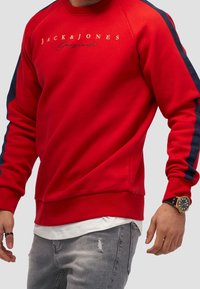 Uomo che indossa un felpa rossa Jack & Jones con strisce blu sulle maniche, jeans strappati grigio chiaro, t-shirt bianca e un orologio da polso nero.