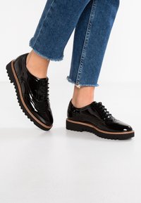 Des chaussures en cuir verni noir présentent un design brogue classique avec fermeture à lacets et une semelle épaisse noire, accentuée d'un bord marron contrastant.